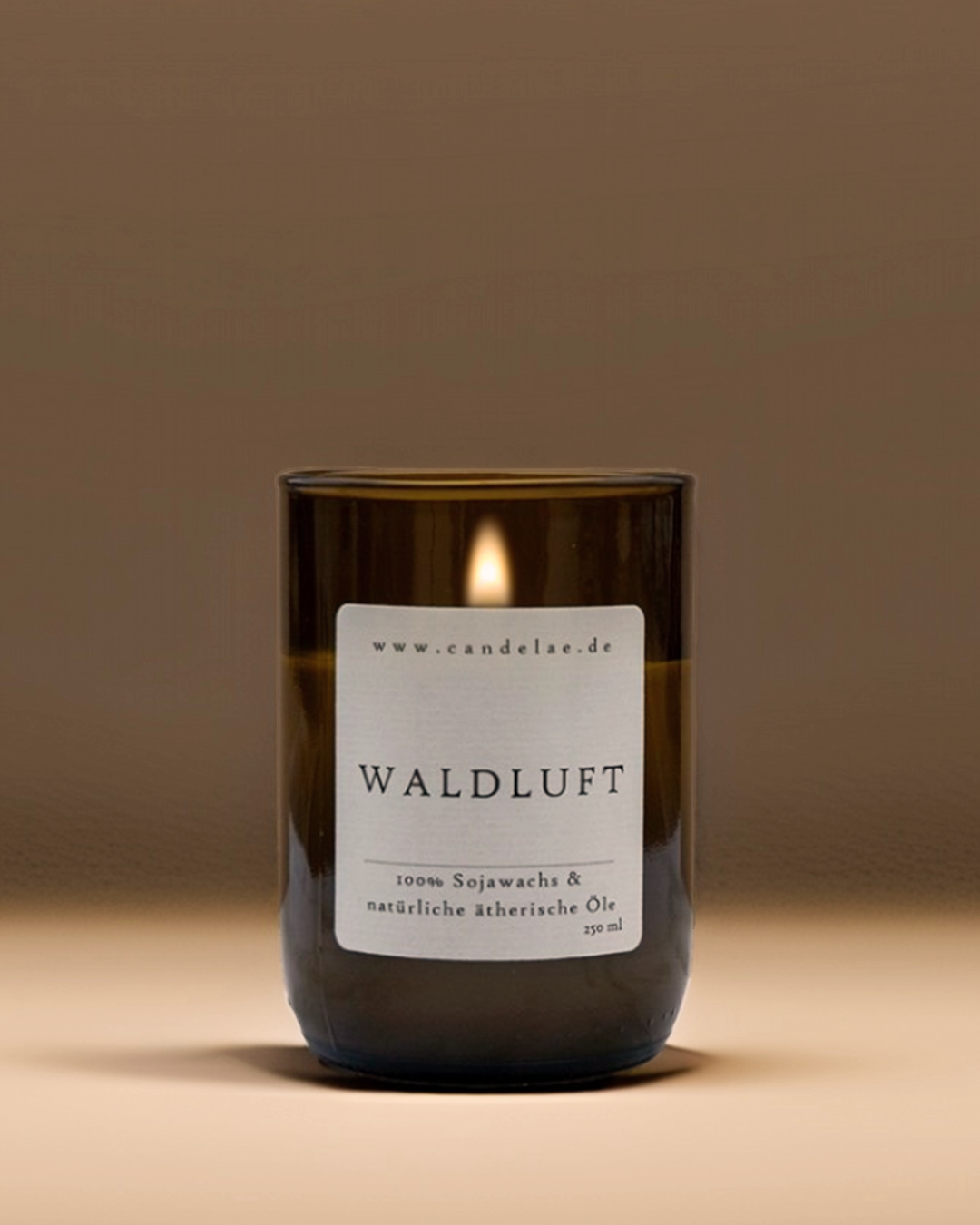 Natürliche Duftkerze „Waldluft“ von candelae in grünem Glas mit brennender Flamme auf beigem Hintergrund – enthält ätherische Öle und Sojawachs.