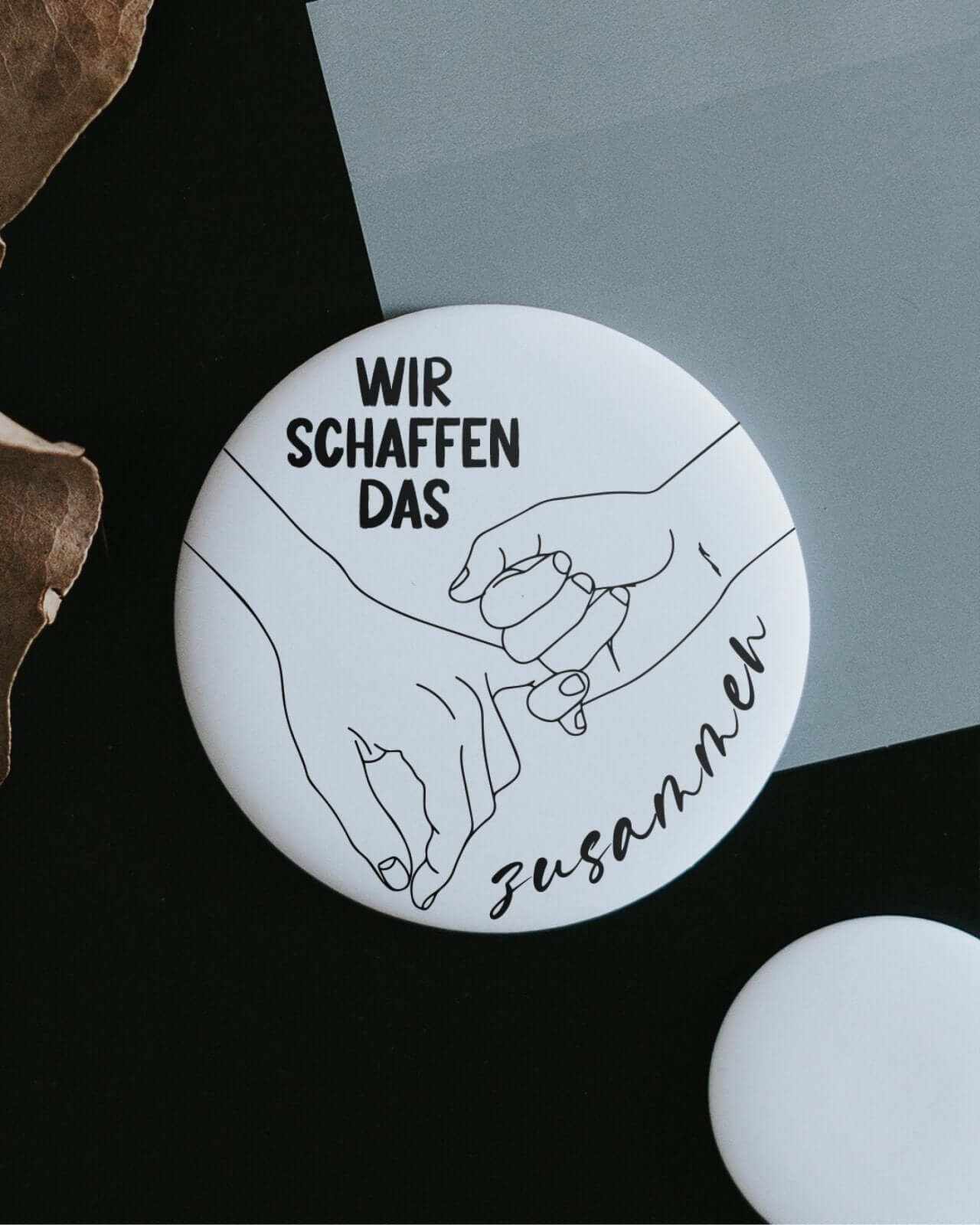 Schwarzer runder Magnet mit weißem Schriftzug „Wir schaffen das zusammen“, symbolisiert Zusammenhalt und Mut