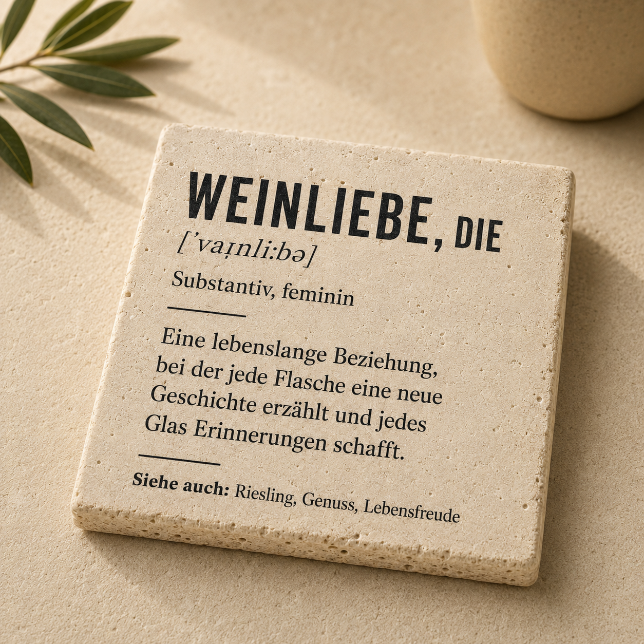 Quadratische Travertinfliese mit schwarzem Weinliebe-Schriftzug und Definition auf hellem Naturstein mit Olivenzweig