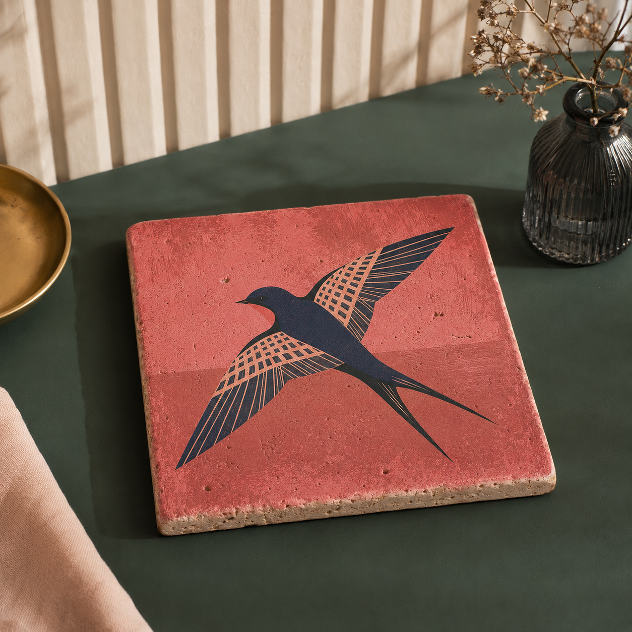 Travertin-Kunstwerk „Höhenflug“ mit fliegender Schwalbe auf korallrotem Hintergrund, liegend auf dunkelgrünem Tisch.