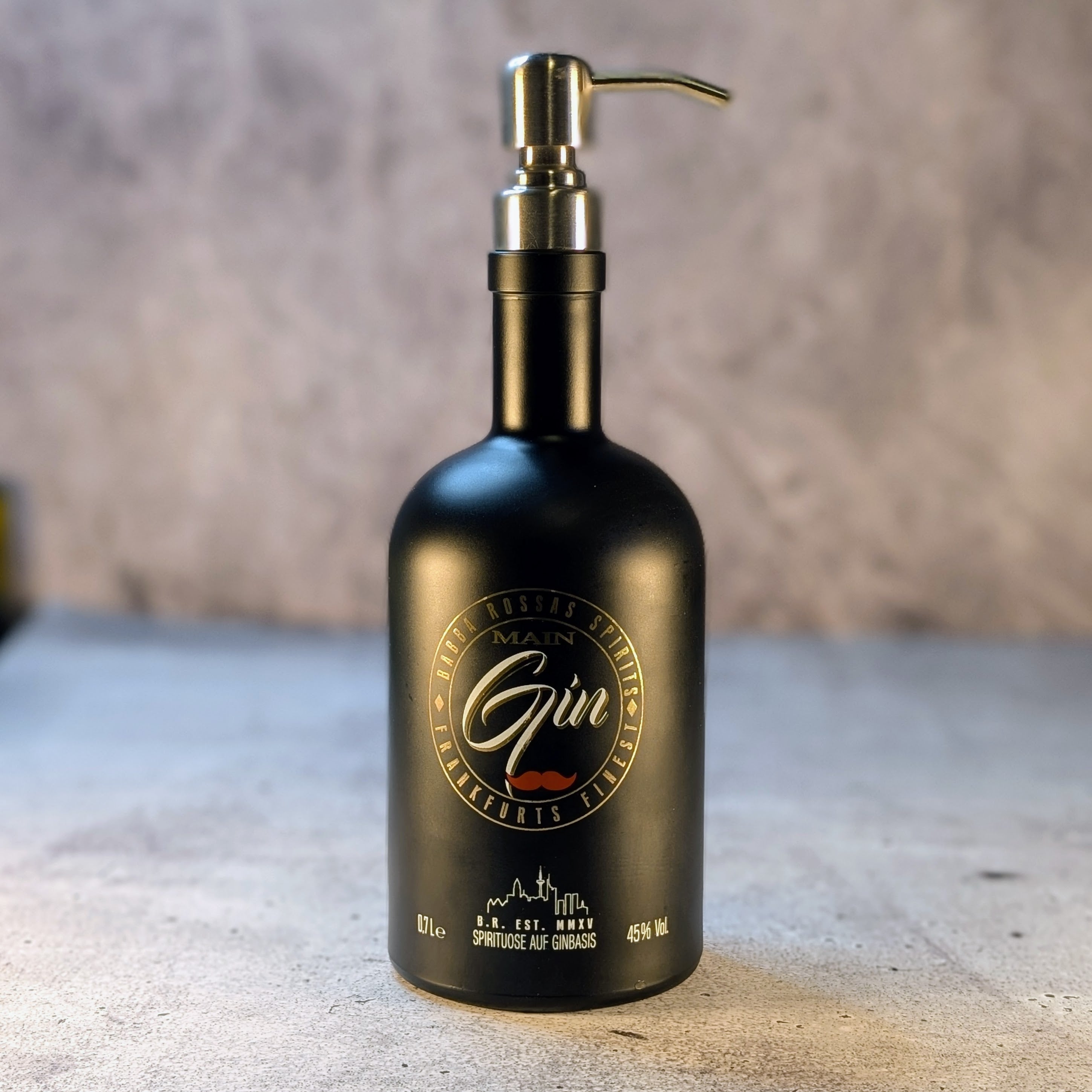 Seifenspender aus einer recycelten "Main Gin"-Flasche mit Pumpenkopf in gebürstetem Silber, stilvoll auf einer hellen Steinoberfläche präsentiert.