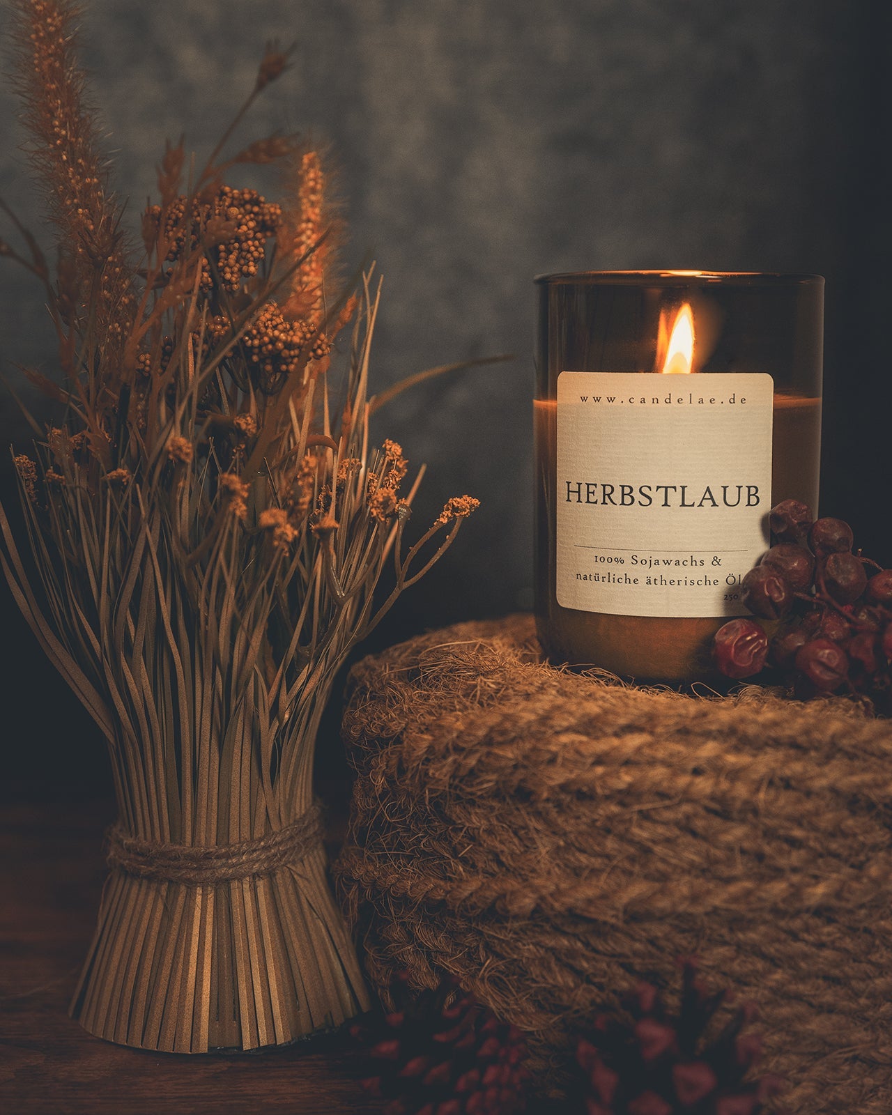 Herbstliche Duftkerze Herbstlaub aus recycelter Weinflasche, brennend neben getrockneten Gräsern und Naturdeko.