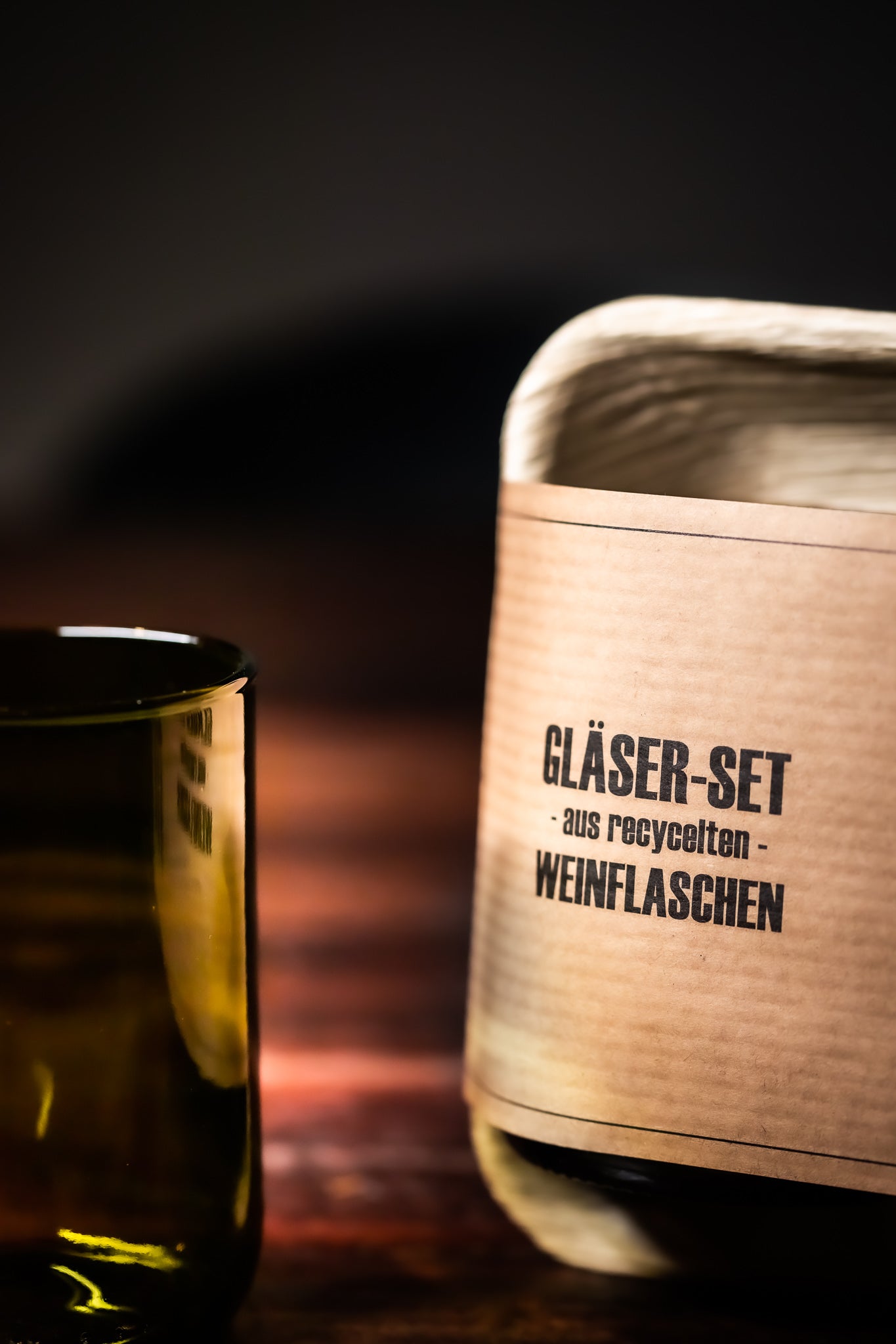 Handgefertigte, nachhaltige Trinkgläser aus recycelten Weinflaschen und umweltfreundliche Duftkerzen, liebevoll arrangiert auf einem rustikalen Holztisch, umgeben von Weinreben und dem Charme Rheinhessens.