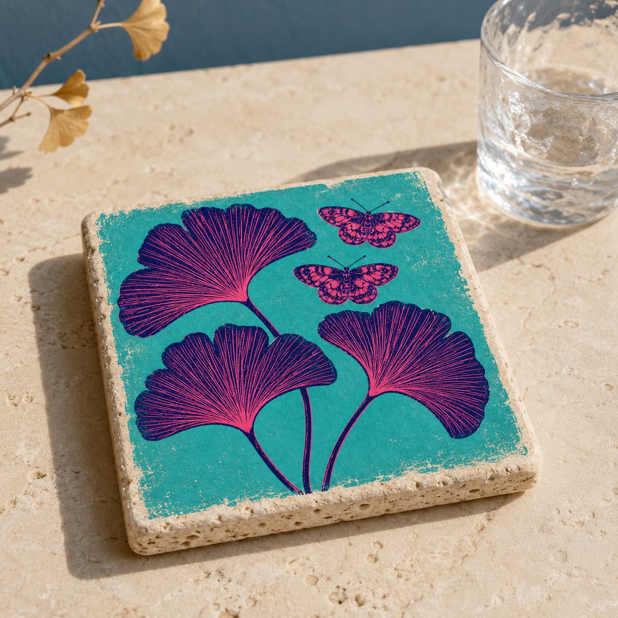 Travertin-Kunstwerk mit pinken Ginkgoblättern und Schmetterlingen auf türkisem Grund neben einem Wasserglas
