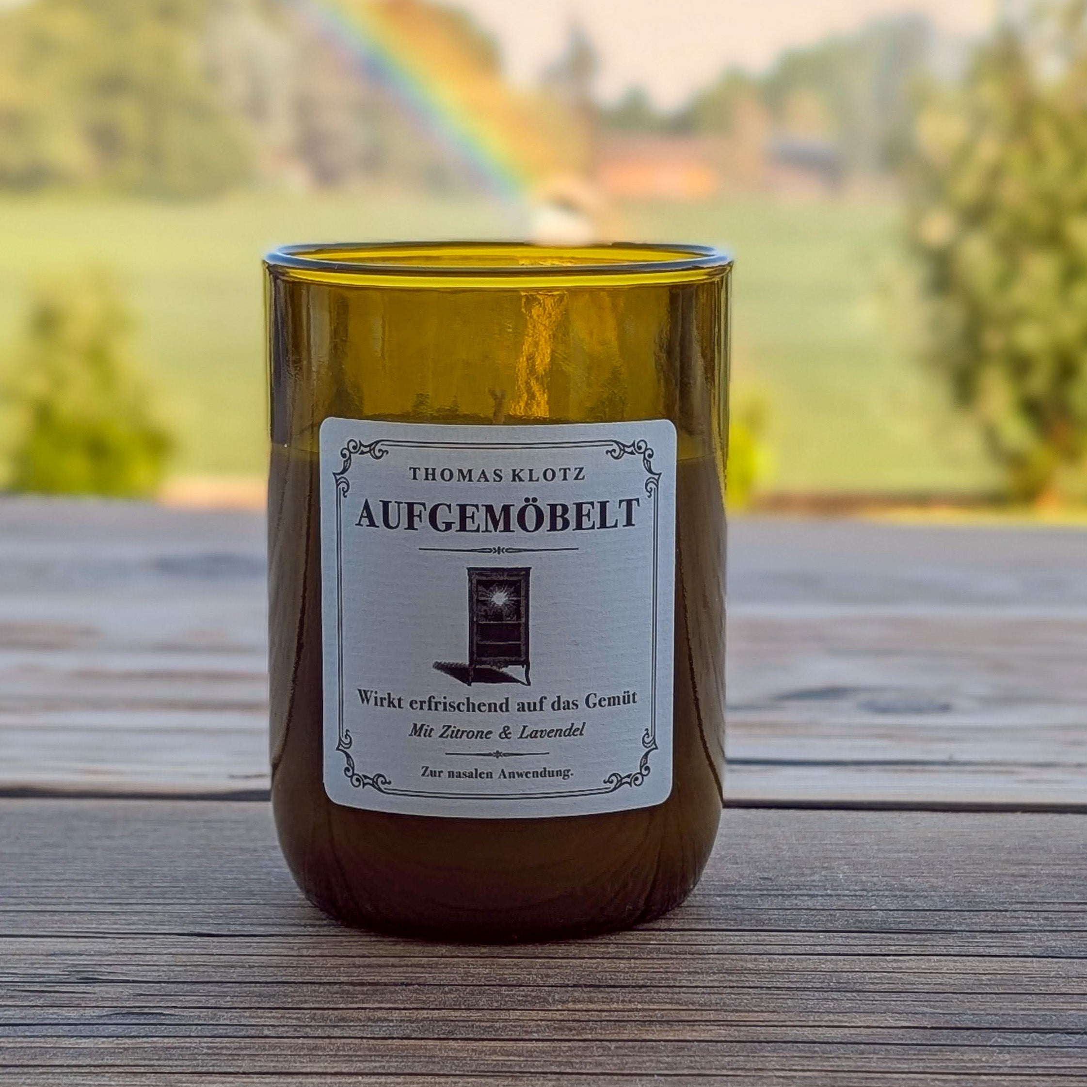 Duftkerze „Aufgemöbelt“ by Thomas Klotz im grünen Recyclingglas – stimmungsvoll inszeniert vor einem Regenbogen auf ländlicher Terrasse