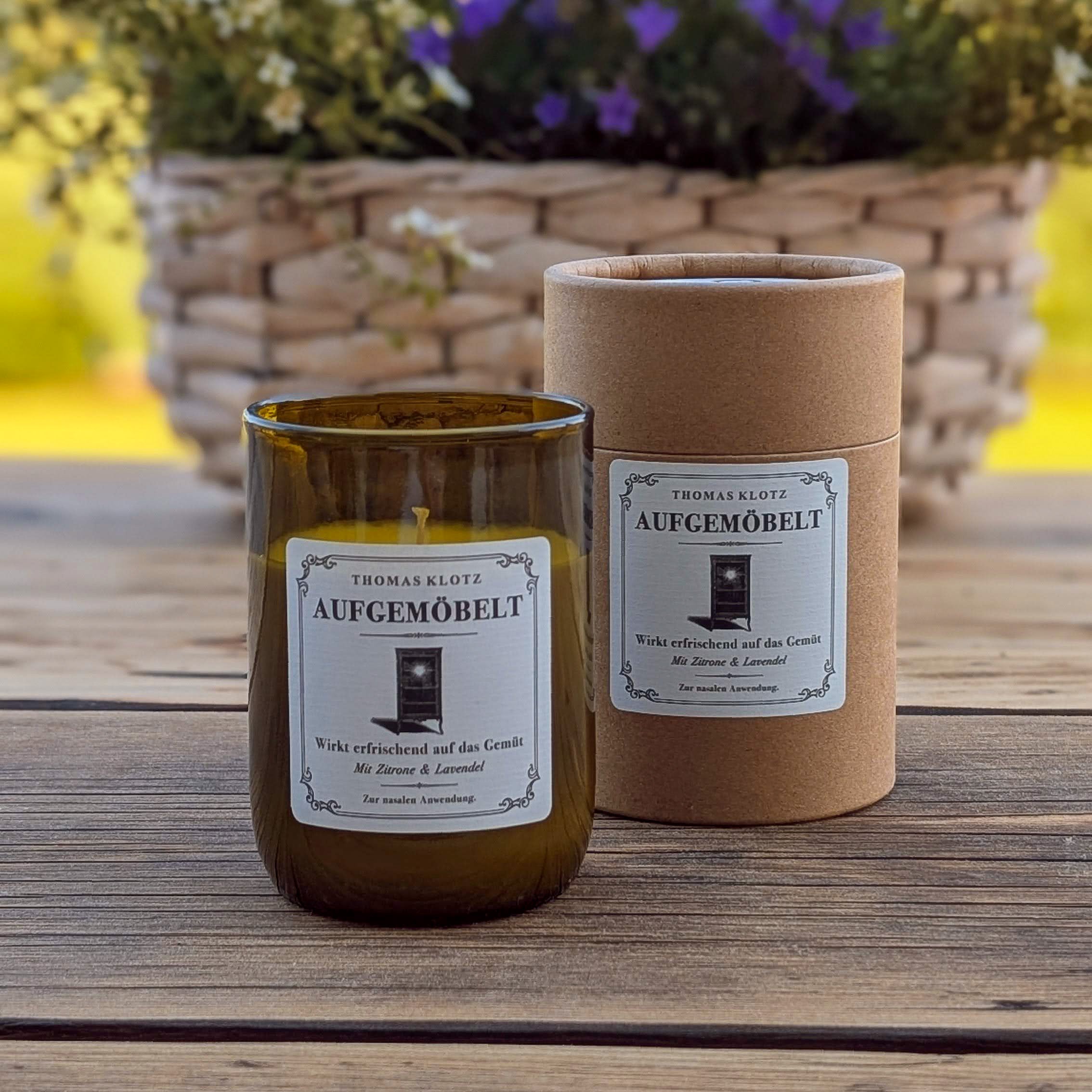 Duftkerze „Aufgemöbelt“ by Thomas Klotz im recycelten Weinglas mit passender Kartonverpackung – präsentiert auf Holztisch mit Blumen im Hintergrund