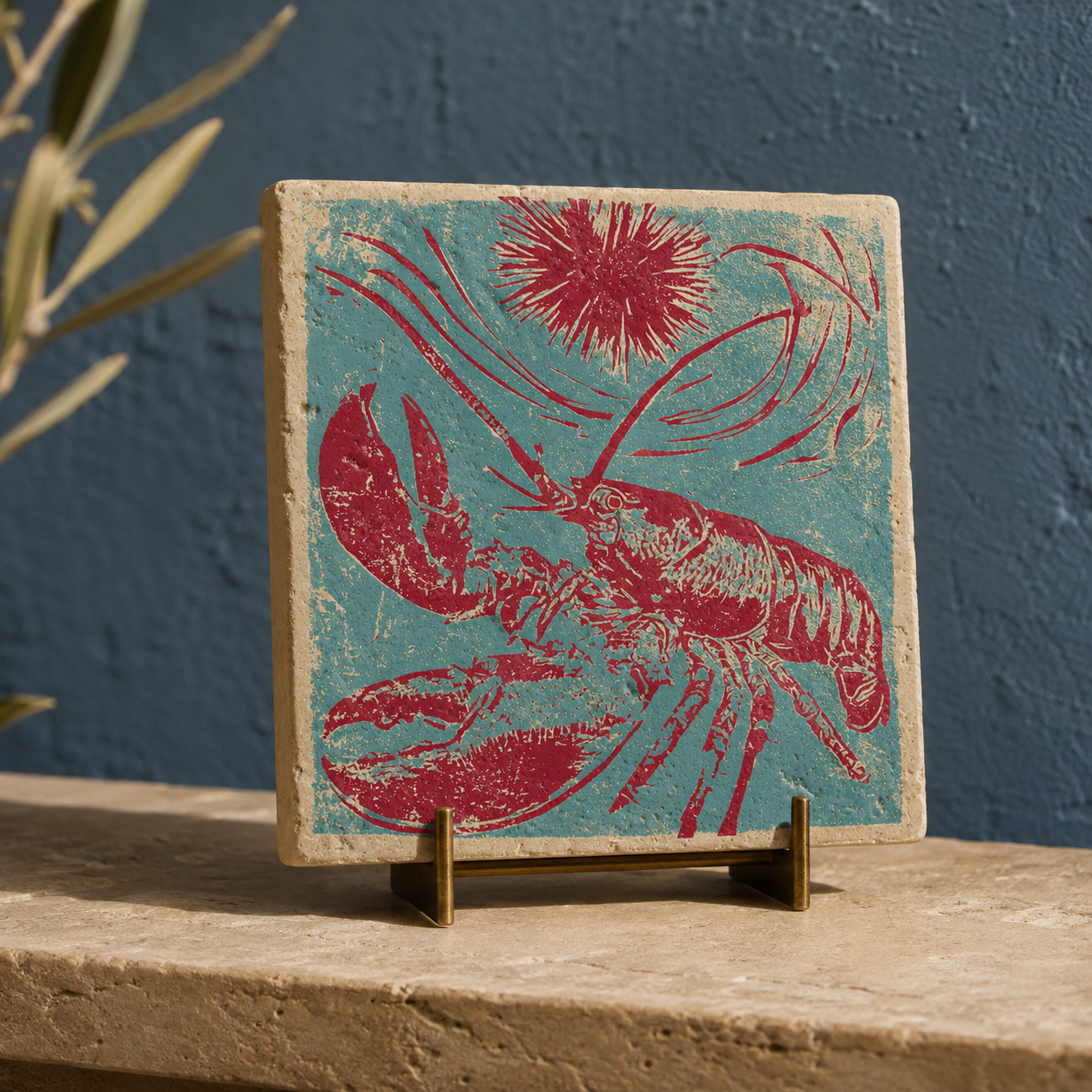 Quadratische Travertin-Dekofliese mit türkis-magentafarbenem Hummer, Seeigel und Wellenlinien auf einem Messingständer. Die 10 × 10 cm große Natursteinfliese steht auf einer hellen Steinkonsole vor einer dunkelblauen Wand.