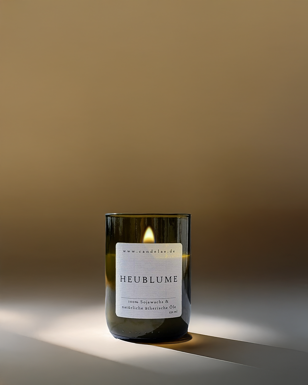 Candelae Duftkerze „Heublume“ – brennend, 250 ml Sojawachs im grünen Glas aus recycelter Weinflasche, mit natürlichen ätherischen Ölen; vegan; warmer, minimalistischer Studiohintergrund.