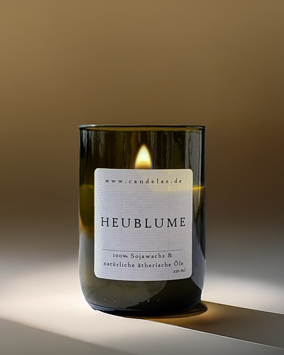 Candelae Duftkerze „Heublume“ – brennend, 250 ml Sojawachs im grünen Glas aus recycelter Weinflasche, mit natürlichen ätherischen Ölen; vegan; warmer, minimalistischer Studiohintergrund.