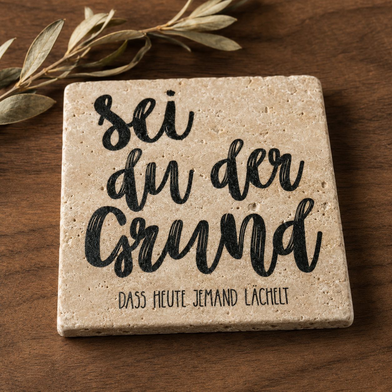 Kleines Kunstwerk aus Travertin mit dem schwarzen Spruch „Sei du der Grund, dass heute jemand lächelt“ auf einem Holztisch mit Olivenzweig.